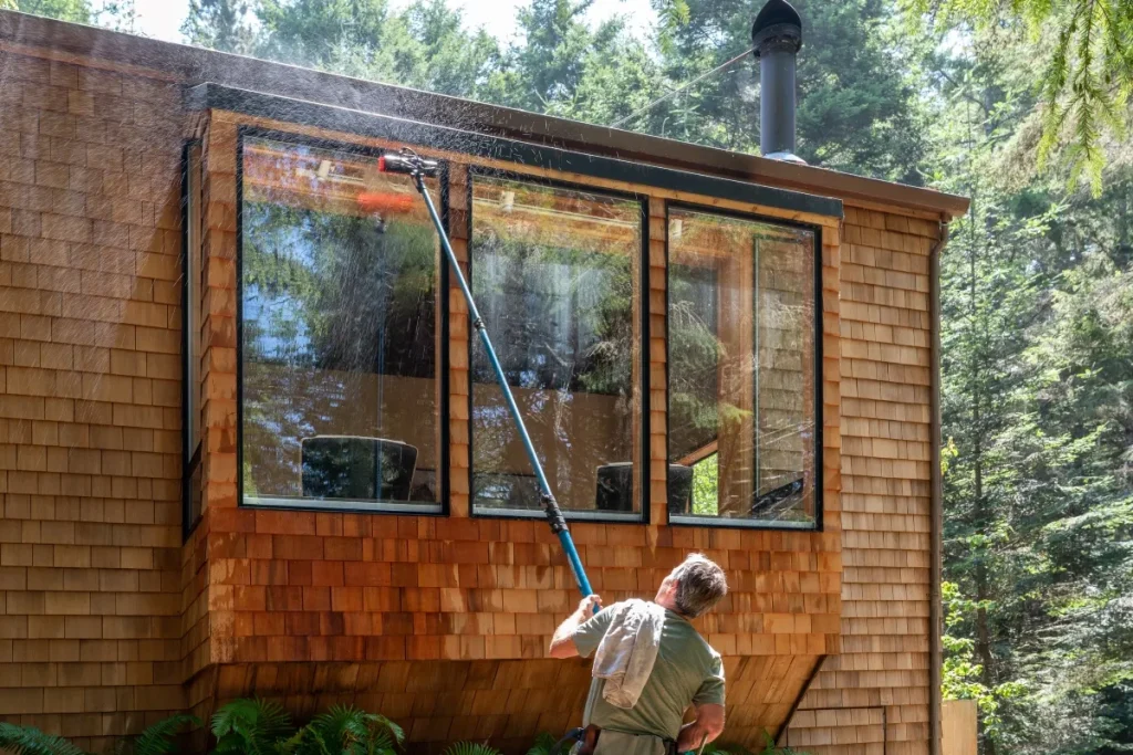 A person cleans large windows of a wooden house using a long pole
