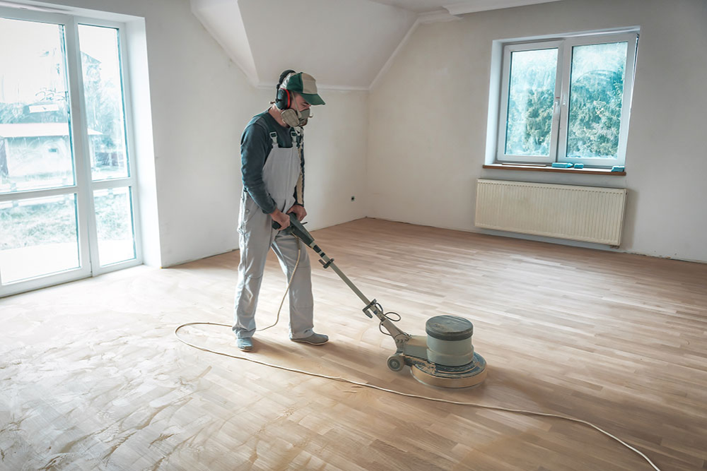 A person sands the wooden floor in a bright, empty room