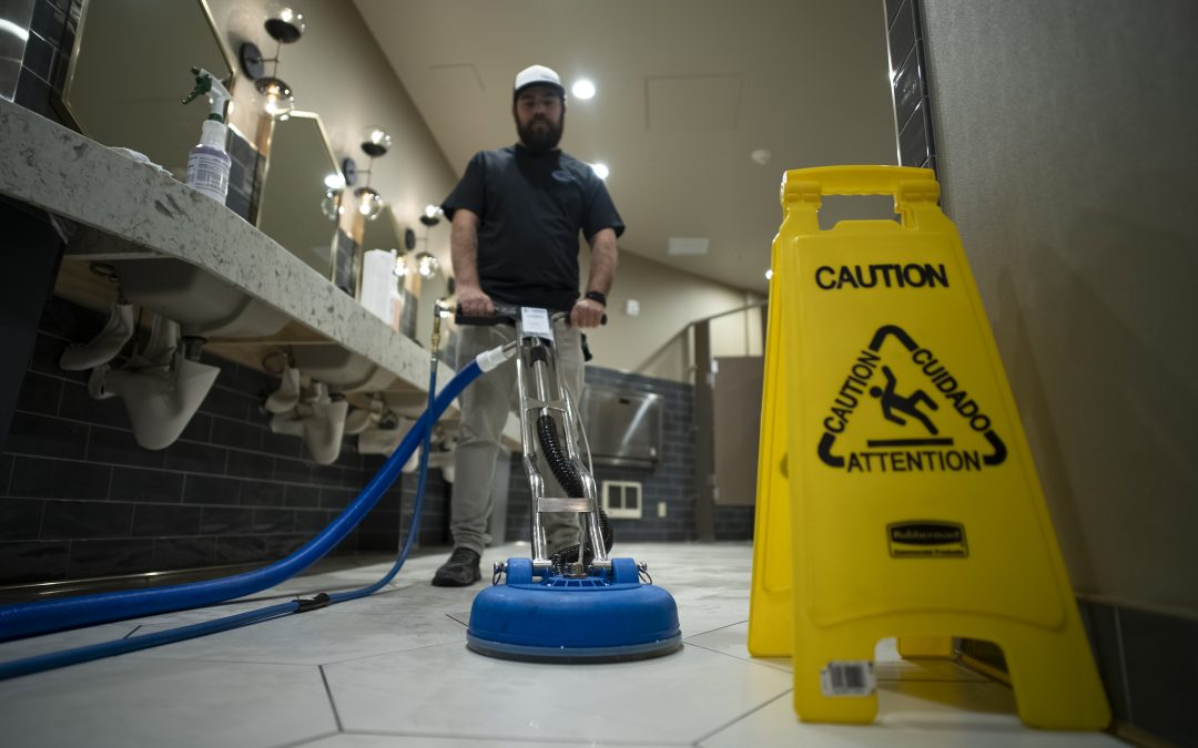 A man operates a floor cleaning machine in a restroom, with a caution sign nearby