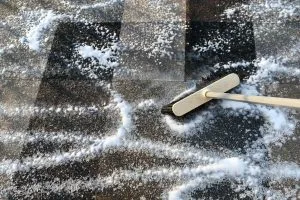 A broom pushes white powder across a dark, textured surface, creating swirling patterns