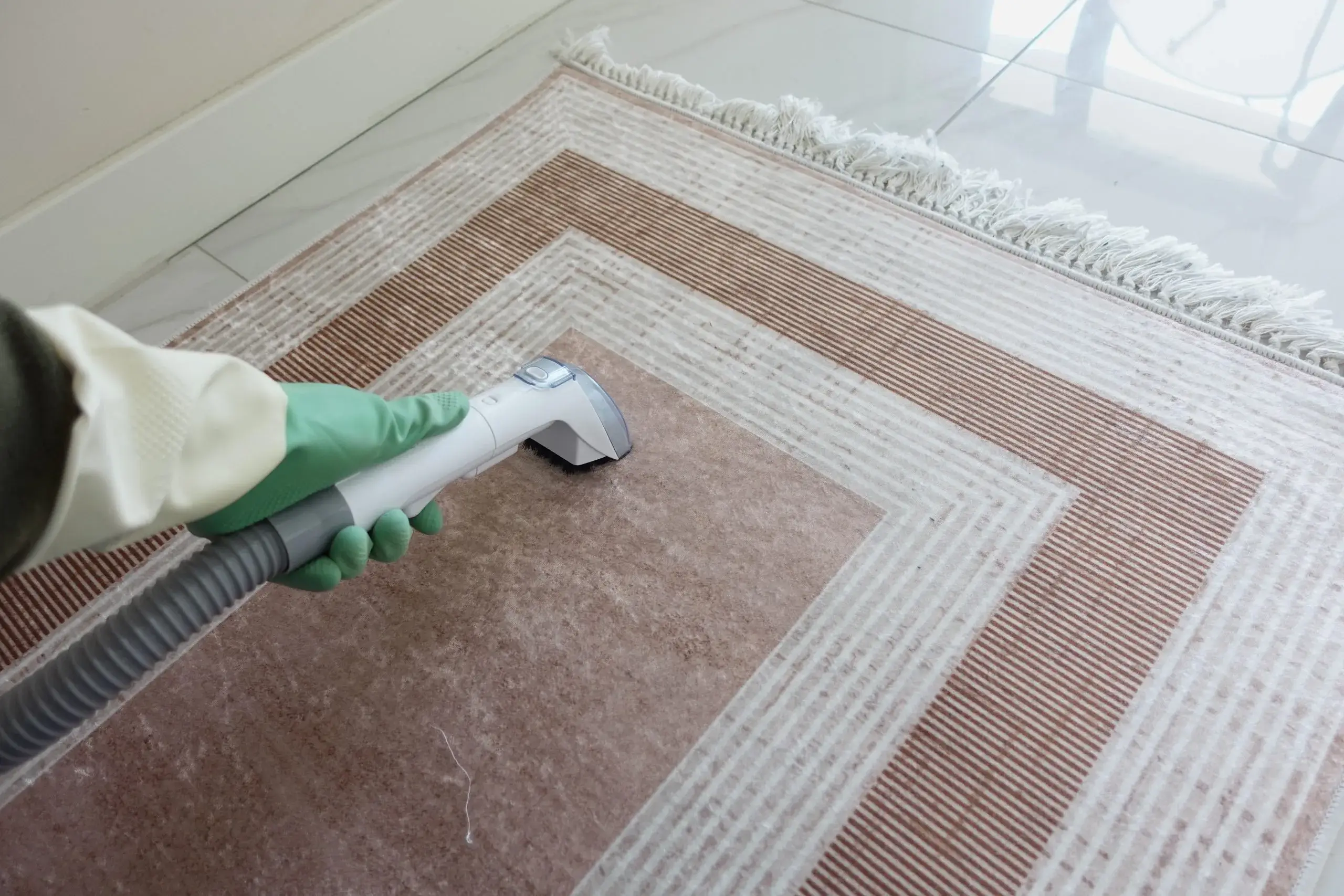 A hand with a green glove uses a vacuum cleaner on a patterned rug
