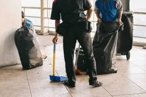 Two individuals clean an area, handling large trash bags and a broom